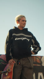 Man wearing a black hoodie with 'Gather' logo holding a skateboard against a graffiti-covered wall.